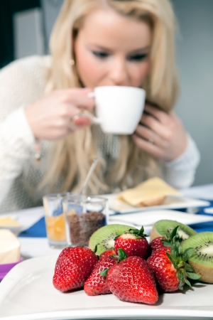 Serving of fresh strawberries and kiwifruit on a white platter on a breakfast table as a healthy appetizer with a blond woman drinking coffee in the background, shallow dofの写真素材