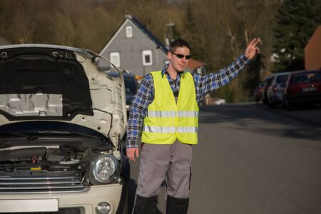 A young man dressed with an reflective vest is standing beside his car and ask for help with his hand.の写真素材
