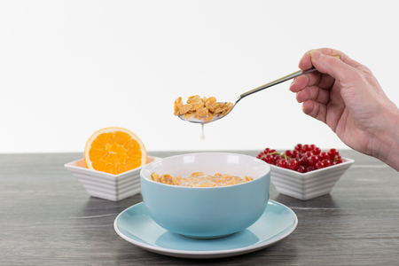 A hand with a spoon of granola and milk. The table is decorated with a orange fruit, a bowl of berry bush and a cup with granola isolated on white.の写真素材