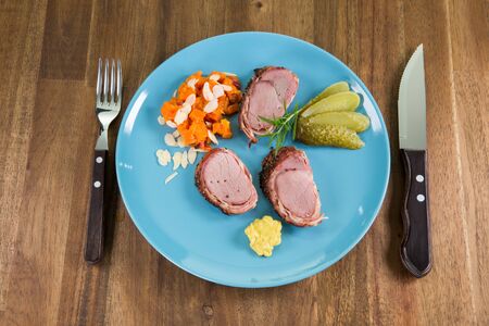 A dish with roast pork, vegetables and mayonnaise on a blueish plate. A fork and a knife besides the plate on a wooden table.の写真素材