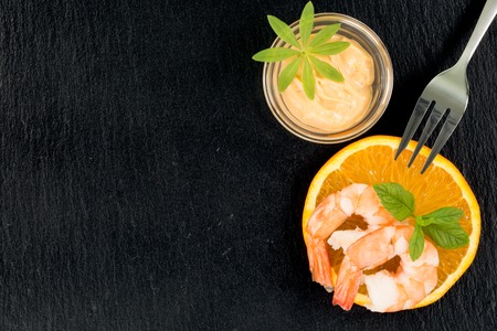 Two big shrimps on a orange fruit isolated on a black slate with copyspace. A small cup of dip decorated with woodruff and a fork on the same side of the plate.の写真素材