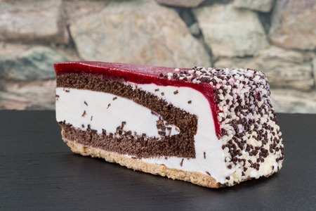 A piece of cherry cream torte on a dark slate with a stone wall in the background.