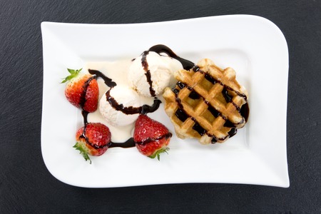 A white plate with waffle, ice and strawberries decorated with chocolate stripes on a dark slate.の写真素材