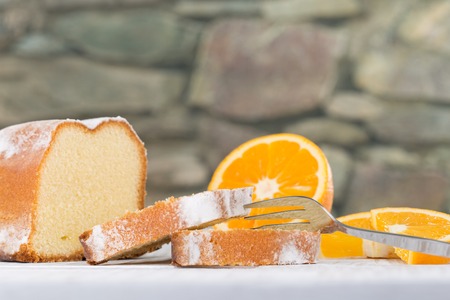A fork beside a sliced lemon cake, decorated with some fresh orange fruit slices. A blurry stone wall in the background.の写真素材