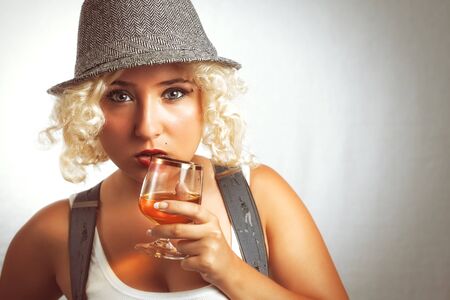 Handsome blonde woman in hat drinking cognac, business style. Young blonde woman with red lipstick. Sexy business woman with glass of whiskey.の写真素材