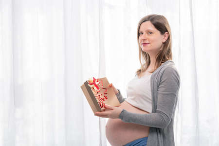 Young pregnant woman with big belly holding a gift wrapped with red ribbon, baby shower party with presents., Gift and happy new year concept.の写真素材