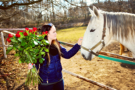 Beautiful young woman standing with bunch of roses before white horseの写真素材