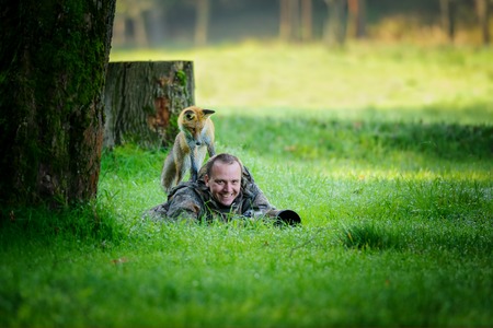 Wildlife photographer hidden in grass with big smile in face, having curious fox on his back looking down to the photographerの写真素材