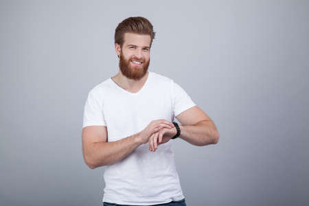 Photo of attractive ginger man with satisfied expression, has thick beard, dressed in casual t shirt, isolated over white studio wall. looking to his watchの写真素材