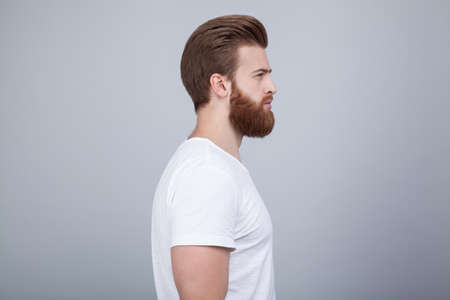 Profile shot of handsome male with trendy hairdo and beard, looks aside with serious expression, has thick red beard, isolated over white backgroundの写真素材