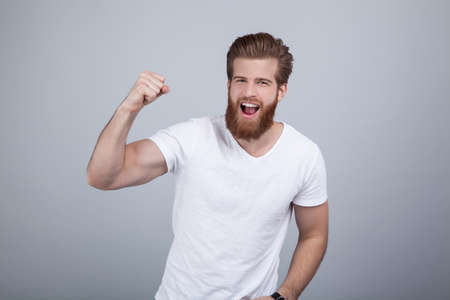 Yes. Winner. Cheerful young man are standing and gesturing for the victory on pure background in casual outfit. Hi is fan of sports games as football, basketball, hockey, baseballの写真素材