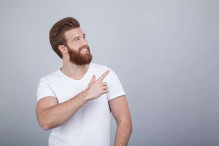 Positive young male freelancer or blogger being satisfied with results of work, has friendly pleasant smile, points upwards with fore finger, wears casual clothes, stands in studio against white wallの写真素材