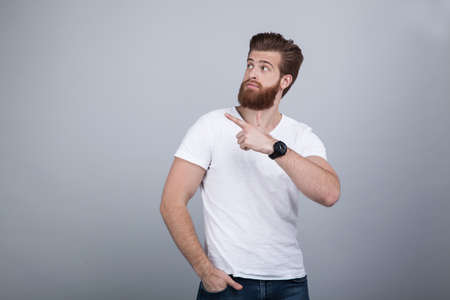 Attractive ginger male has thick beard, dressed in casual outfit, has serious expression, points with index finger at upper right corner, shows free space for your advertisement or promotional textの写真素材