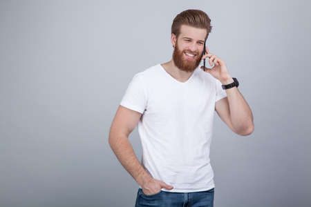Happy handsome bearded man in white t shirt talking on cell phone over white backgrounの写真素材