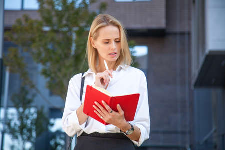 thoughtful blonde business woman with notebook against of office buildingの写真素材