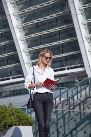 Happy blonde business woman in sunglasses with notebook against of modern buildingの写真素材