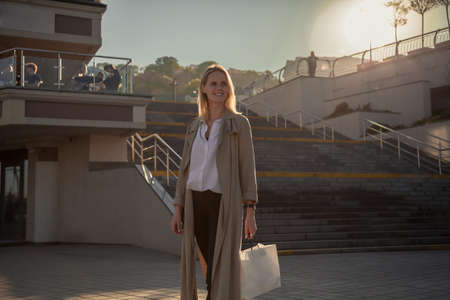 Young woman in a stylish spring coat in fashionable glasses in a white T-shirt in a black leather handbag walking along the street against the background of vintage buildings. European girl.の写真素材
