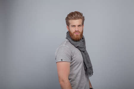 young handsome bearded man standing by the grey wall wearing scarf and grey t-shirt rotated in profile with her head turned to the camera.の写真素材