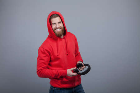 young handsome smiling bearded man in red hoodie on holding headphones in his hands standing at the grey background and looking to the camera.の写真素材