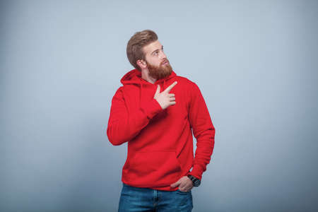 young handsome bearded man wearing red sweatshirt standing on the grey background holding his hand in the pocket and pointing his finger on the right side and looking there with the interest.の写真素材