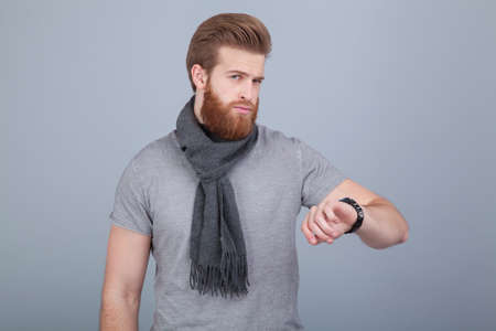 young handsome bearded man wearing scarf standing by the grey wall looking at his watch checking the time.の写真素材
