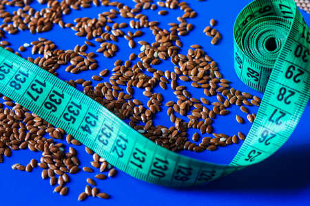 brown flax seeds are scattered on a solid, bright blue background next to a twisted centimeter of green flower. The topic of losing weight with flax seeds and cleansing the bodyの写真素材