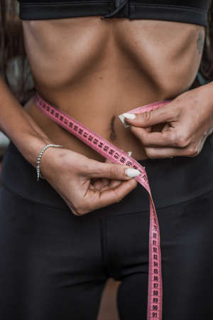 a slender dark-skinned girl in a sports uniform pulls in her stomach and measures the waist with a centimeterの写真素材