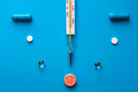 A mercury thermometer for measuring body temperature, surrounded by multi-colored tablets and capsules, shows vitamin C in the form of an orange orange. Thermometer and pills are laid out on a blueの写真素材