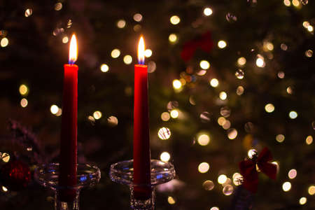 Two long red candles in tall glass candlesticks against the background of the lights of the New Year tree. A candlelit celebration in a romantic setting. Bokeh effect in the backgroundの写真素材