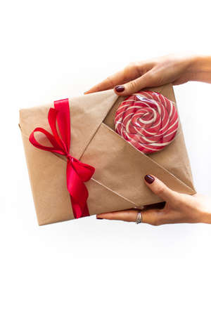 Beautifully wrapped gift in kraft paper with a red bow and a bright sweet candy for decoration in the hands of a girl. The girl holds a gift in her hands, photo on a white clean background, top view. The girl gives a gift. The girl accepts a gift. Banner with place for text. High quality photoの写真素材
