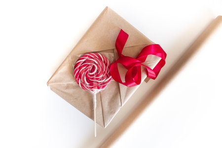 Holiday gift wrapping. A box wrapped in beige craft paper lies next to a red ribbon and a round lollipop candy for decoration on a white background. Valentines Day Gift Wrapping Processの写真素材