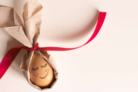 Beautiful Easter egg with a painted smile, eyelashes and freckles in bunny ears from a napkin on a white background. Easter Ukraine 2022. Cool egg for Easter with a painted face and a red bright ribbon. High quality photoの写真素材