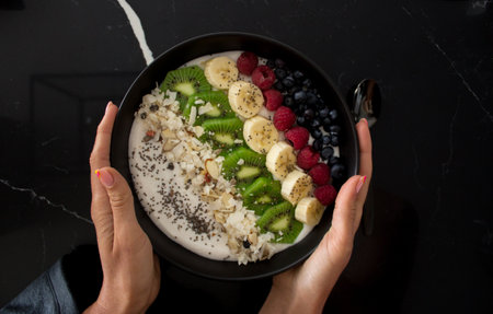 Female hands hold a black plate of oatmeal with fresh fruits, nuts and chia seeds, top view, healthy fast food. Useful healthy breakfast. Slimming. Beautiful presentation of the food.の写真素材
