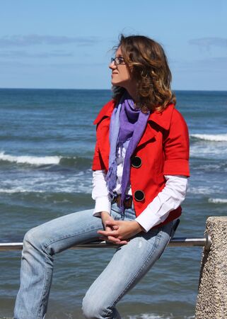 Beautiful fashionable girl sitting on the railing in front of the seaの写真素材