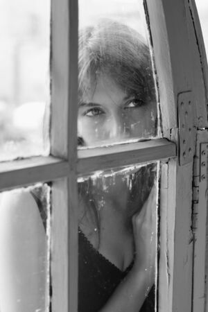 Art portrait of a beautiful young sadly girl looking through the windowの写真素材