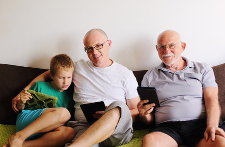 father, grandfather and son playing games on tablets and reading ebookの写真素材