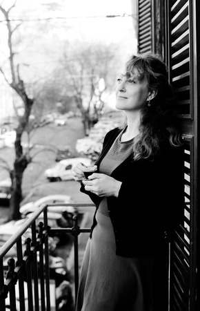 Beautiful mature woman standing on the balcony with coffee looking at the streetの写真素材
