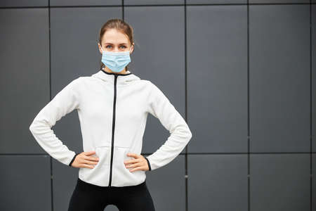 Portrait of a young beautiful woman in a white sports jacket and mask standing on a grey city background after running on the streets. Sport and healty style.の写真素材