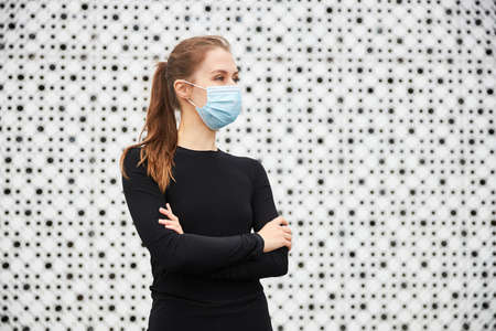 A young girl stands in the cityscape opposite a modern building with a white textured wall and looks into the distance dressed in a black sports jacket and a medical mask. Empty space for your text.の写真素材