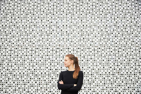 A young girl stands in the cityscape opposite a modern building with a white textured wall and looks into the distance dressed in a black sports jacket. Empty space for your text.の写真素材