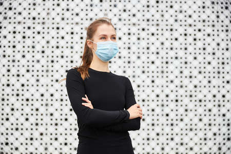 A young girl stands in the cityscape opposite a modern building with a white textured wall and looks into the distance dressed in a black sports jacket and a medical mask. Empty space for your text.の写真素材