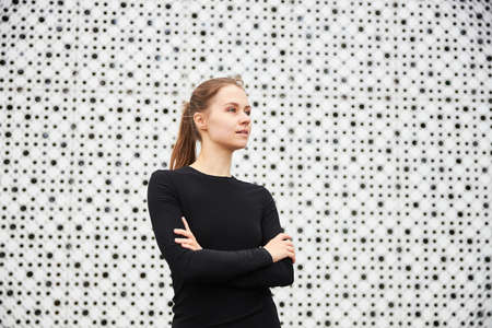 A young girl stands in the cityscape opposite a modern building with a white textured wall and looks into the distance dressed in a black sports jacket. Empty space for your text.の写真素材