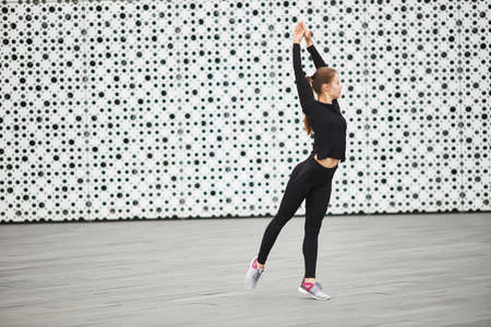 A young girl trains on the street in a black tracksuit. The white textured wall of the city building in the background. Empty space for your text.の写真素材