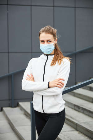 Portrait of a young beautiful woman in a white sports jacket and mask standing on a grey city background after running on the streets. Sport and healty style.の写真素材