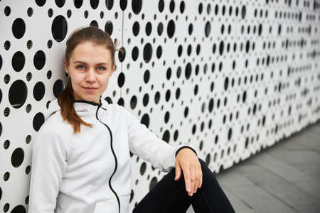 A girl in a white tracksuit rests after a sport near the city wall.の写真素材