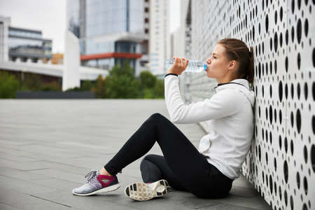 A girl rests and drinks water after sports in the city.の写真素材