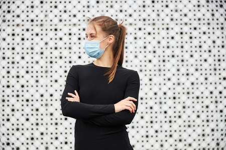 A girl in a black tracksuit and a medical mask stands in front of a white city wall. A place for text.の写真素材
