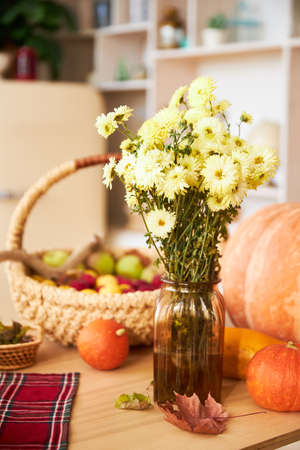 Different multicolored decorative pumpkins, wooden plates or dishes and vase with yellow chrysanthemums flowers on a wooden table on a background of white wall. Autumn or fall home interior decor.の写真素材