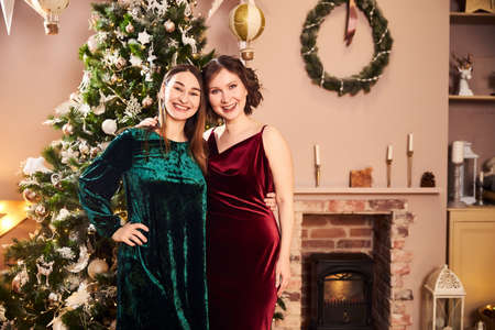 Two beautiful women in eternal dresses have arranged a party for the occasion of the New Year or Christmas in a beautiful room decorated with a Christmas tree and garlands and gifts near the fireplaceの写真素材