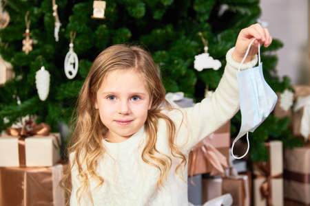 A little cute girl holds a medical mask, a Christmas tree and presents at the backの写真素材